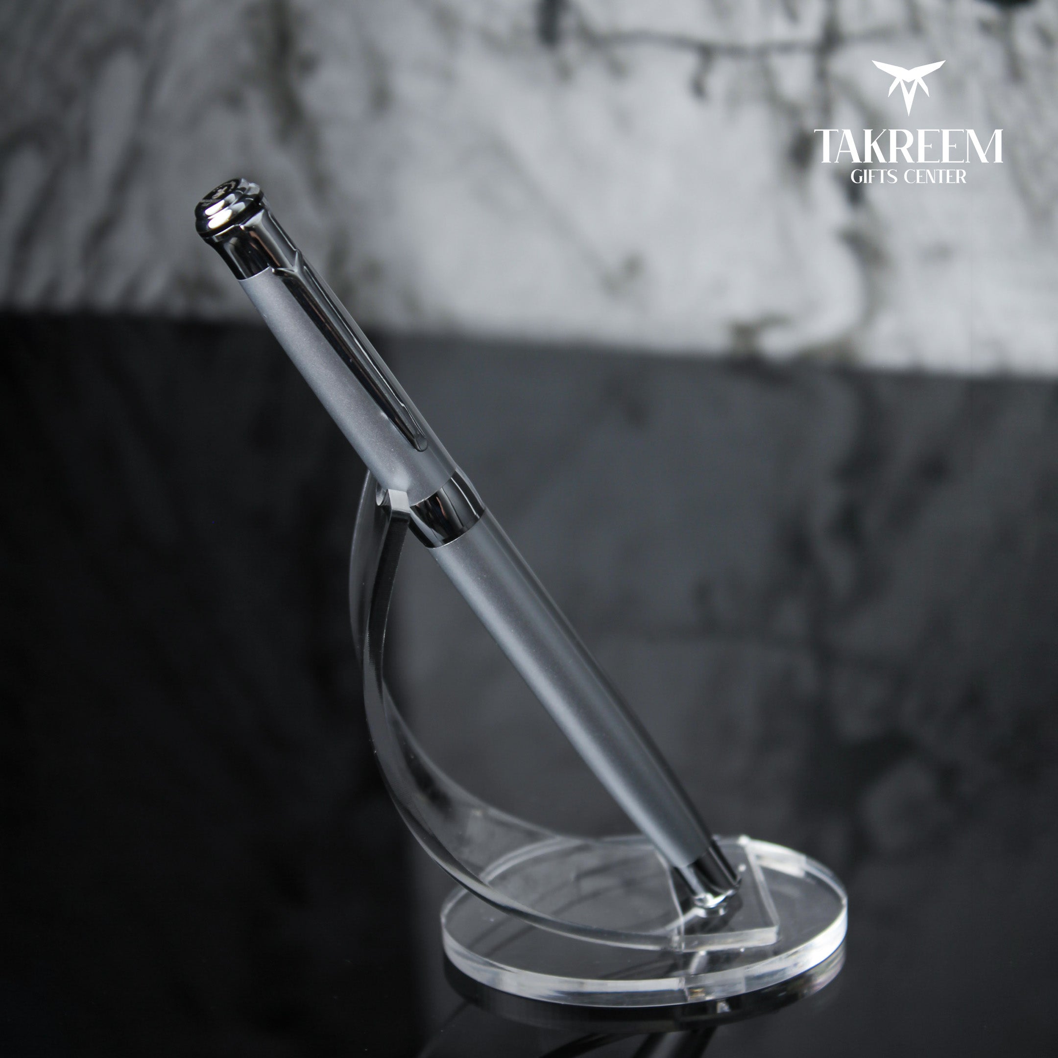 Silver pen on a clear stand with a blurred background and 'Takreem Gifts Center' logo.