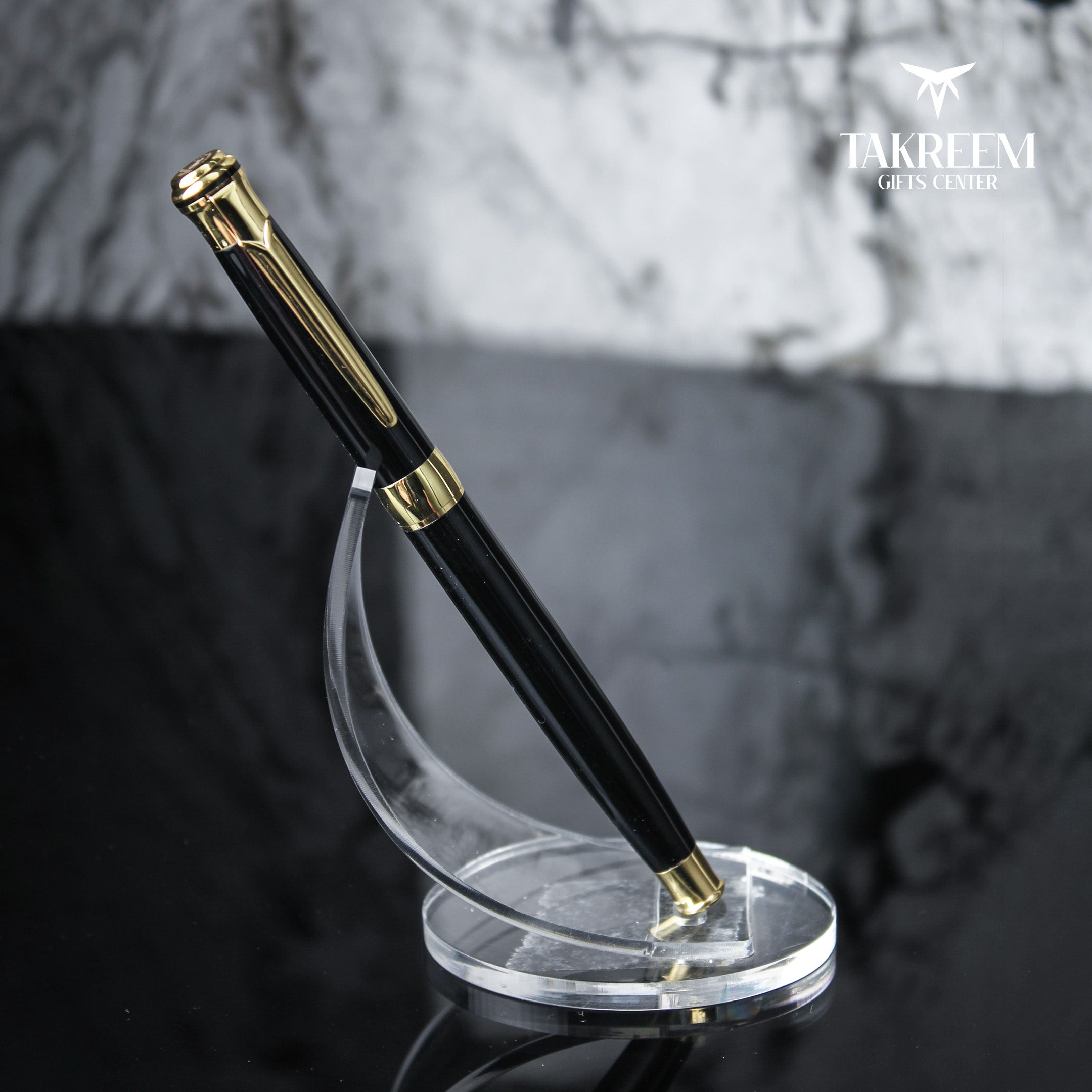 Black pen with gold accents on a clear stand against a marble background, featuring the Takreem Gifts Center logo.