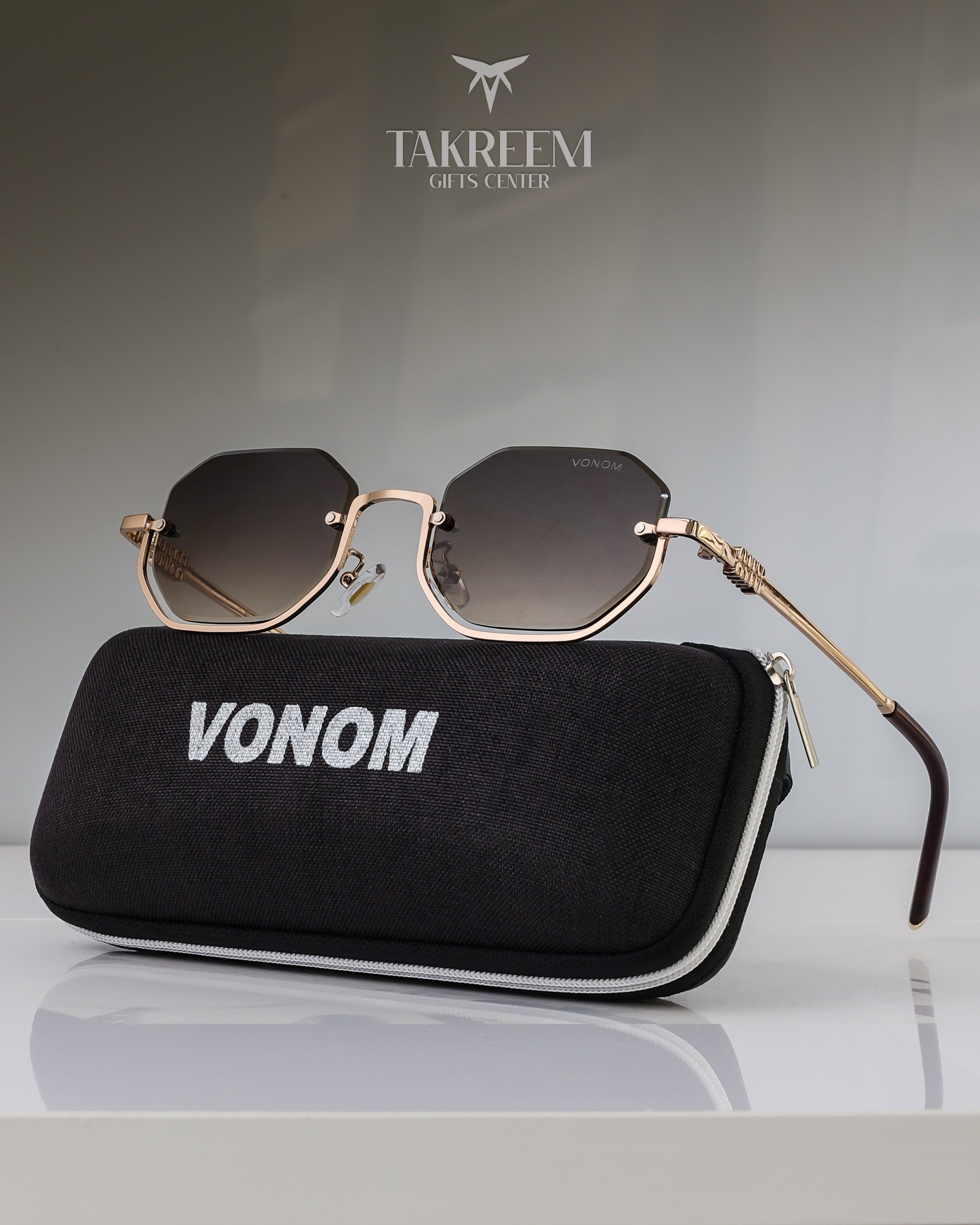 Sunglasses with gold frame and black lenses on a black case labeled 'VONOM' with 'Takreem Gifts Center' logo in the background.