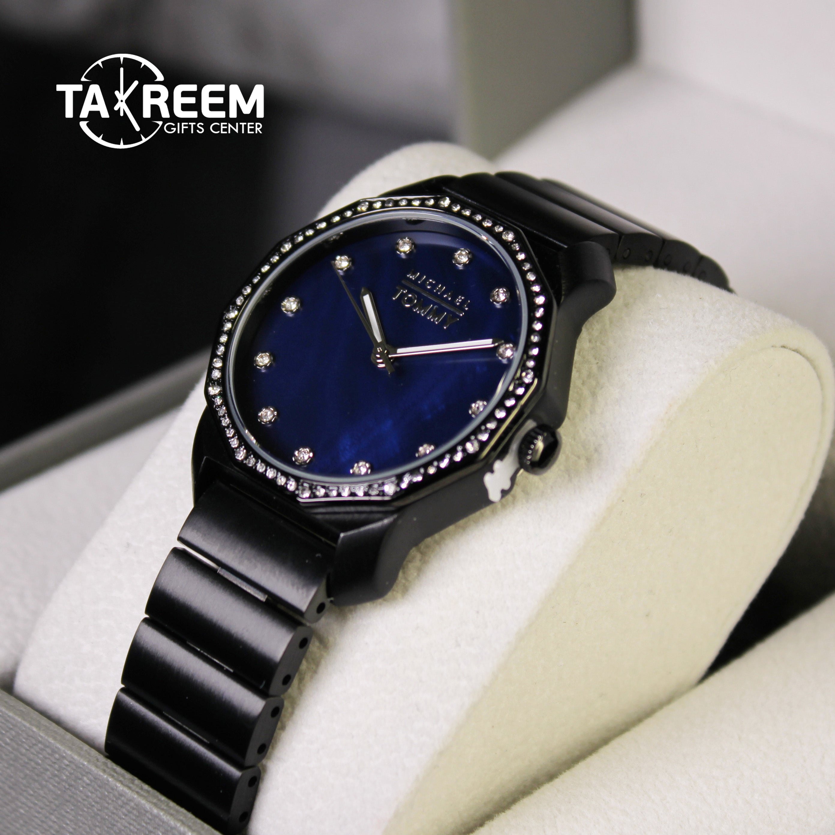 Black wristwatch with blue face and diamond-like stones on a white cushion, brand 'Takreem' visible.