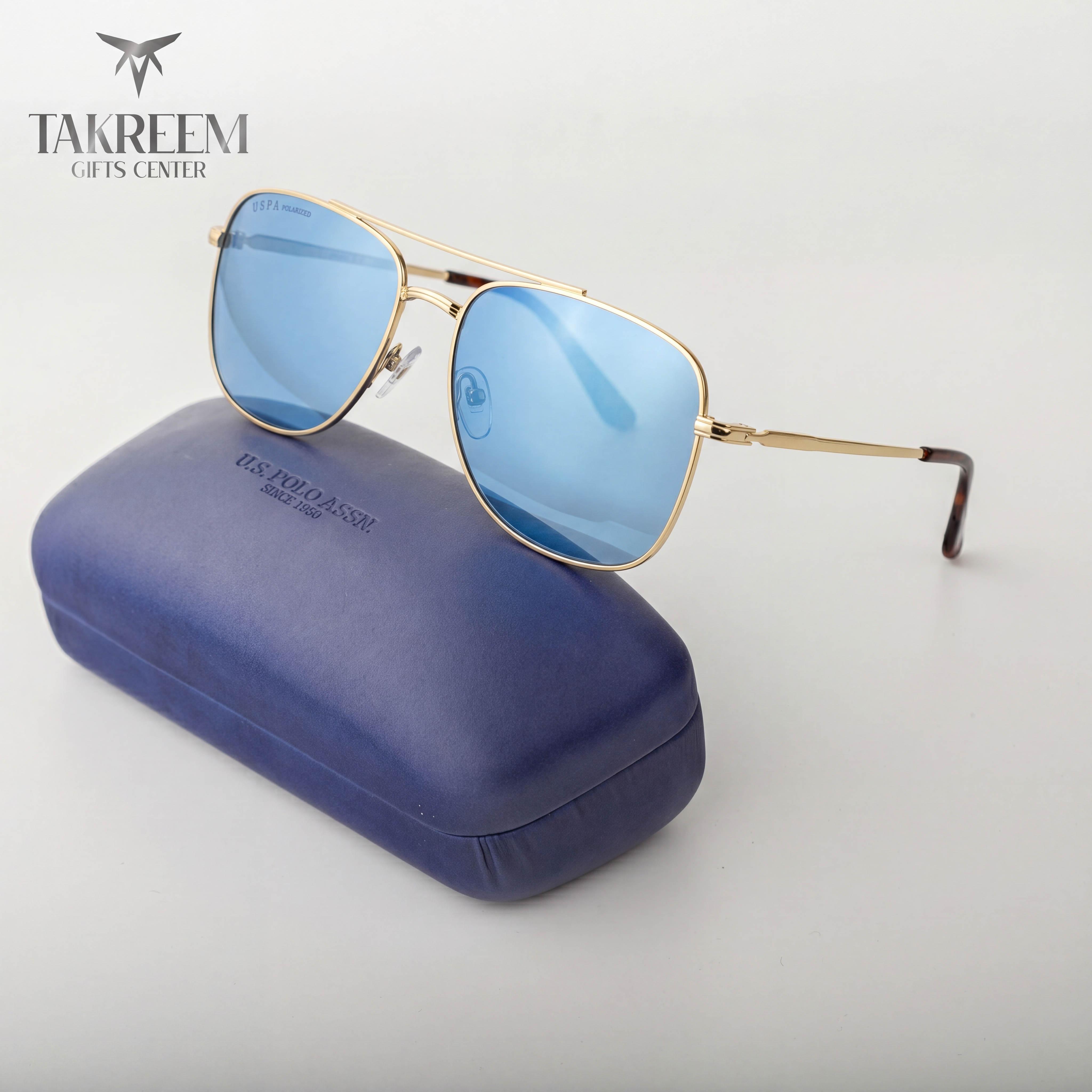Gold-framed sunglasses with blue lenses on a blue case, branded 'Takreem Gifts Center'.