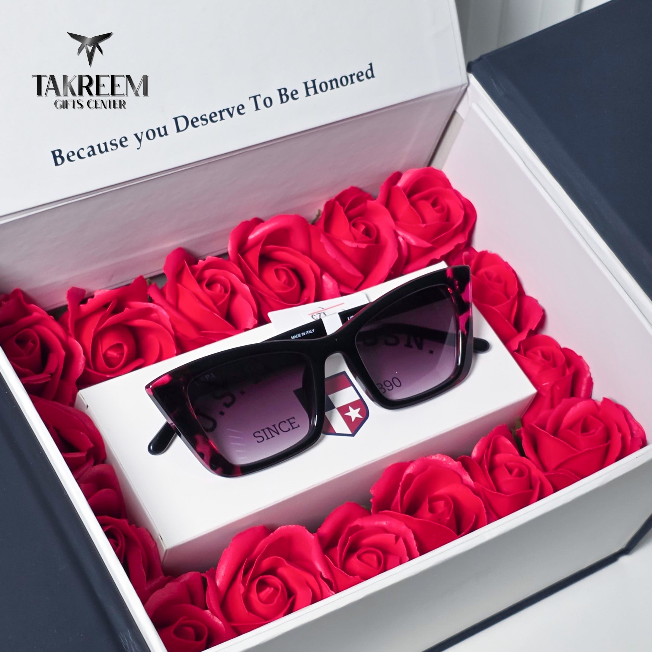 Black square sunglasses with dark lenses resting on a white branded box inside an open presentation box filled with red roses. The inner lid shows the Takreem Gifts Center branding.