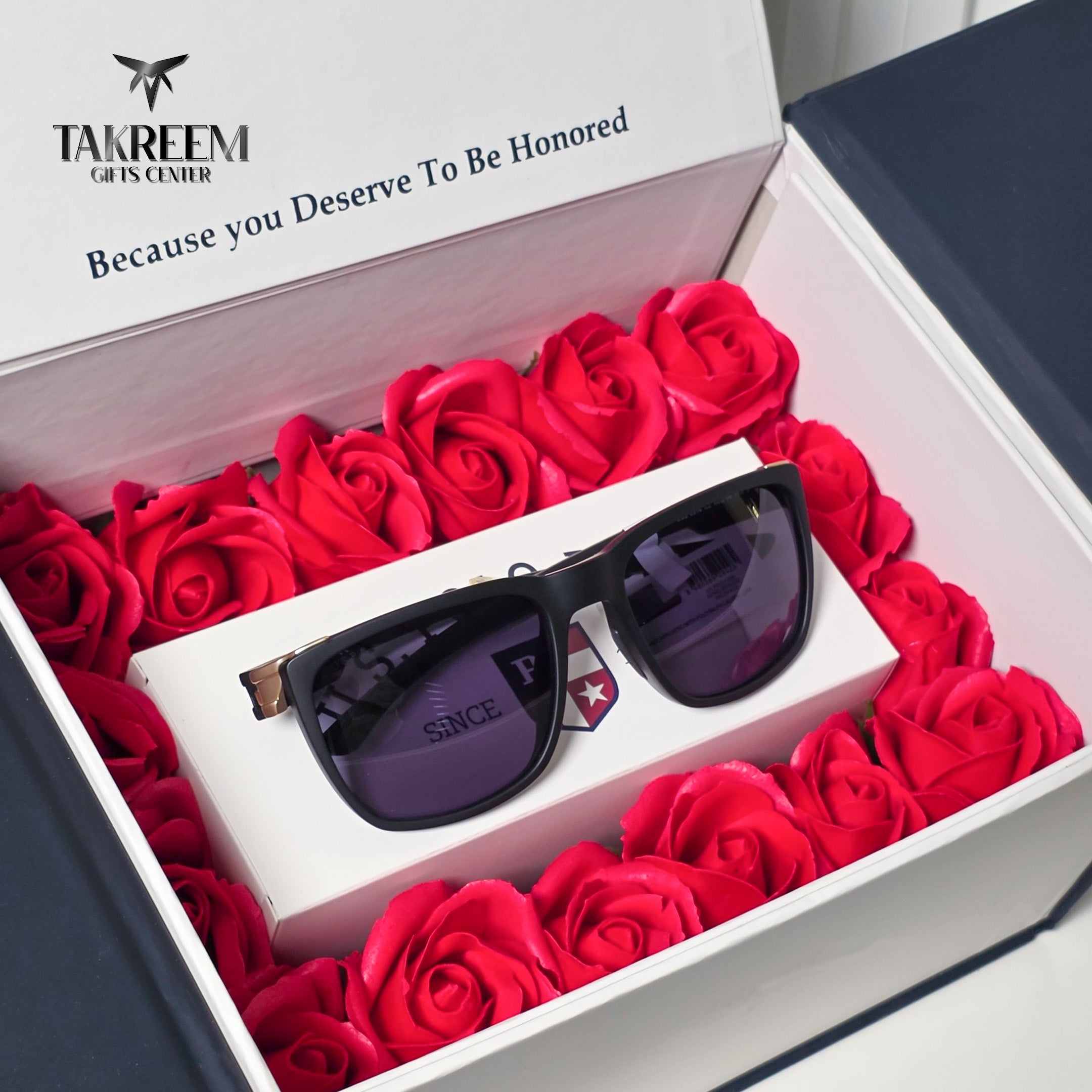 Black square sunglasses with dark lenses resting on a white gift box inside an open presentation box filled with red roses. The inner lid displays the Takreem Gifts Center logo and slogan.