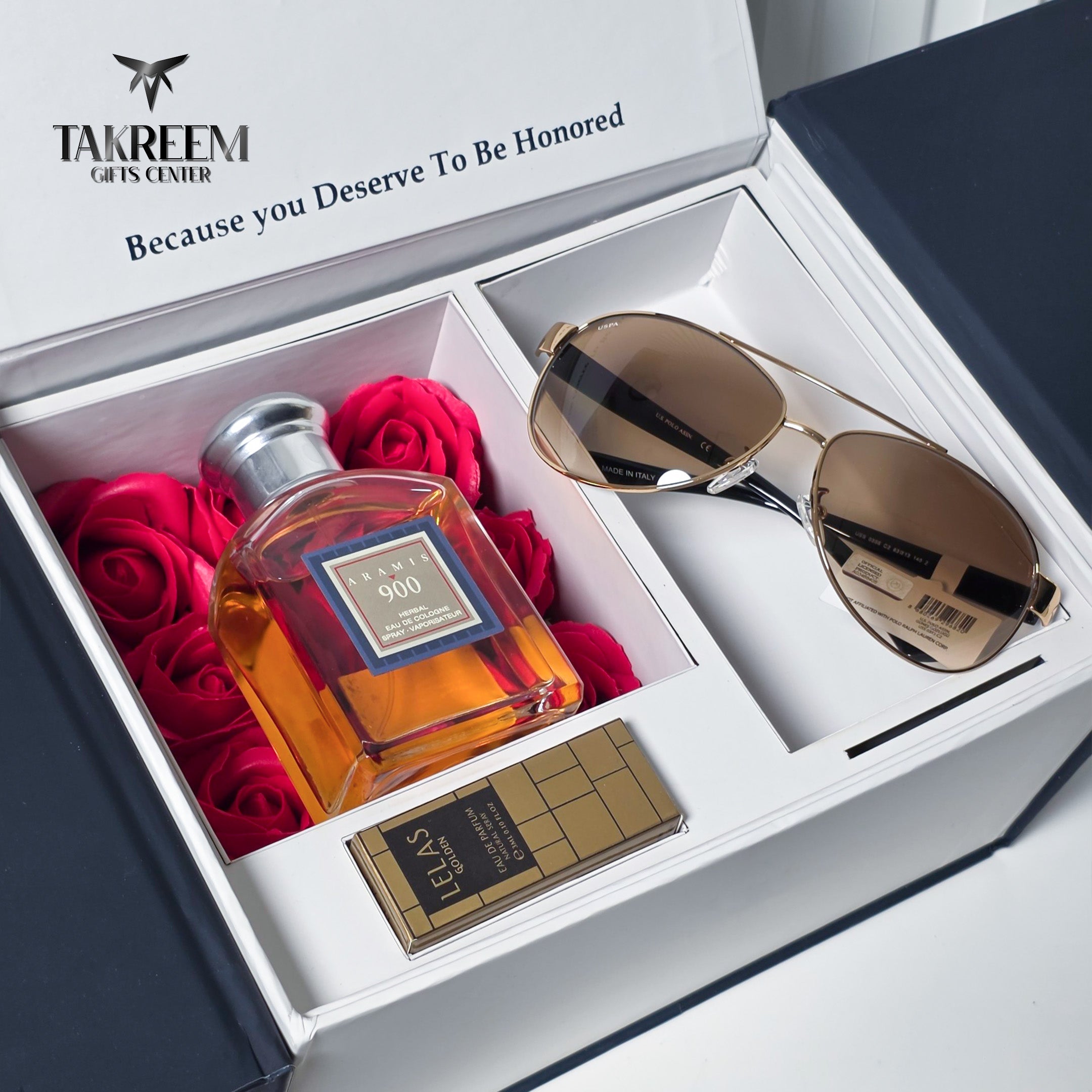 A luxury gift set from Takreem Gifts Center, featuring a bottle of Fram 900 cognac, gold aviator sunglasses, and a chocolate bar, all arranged inside an open white presentation box filled with red roses.