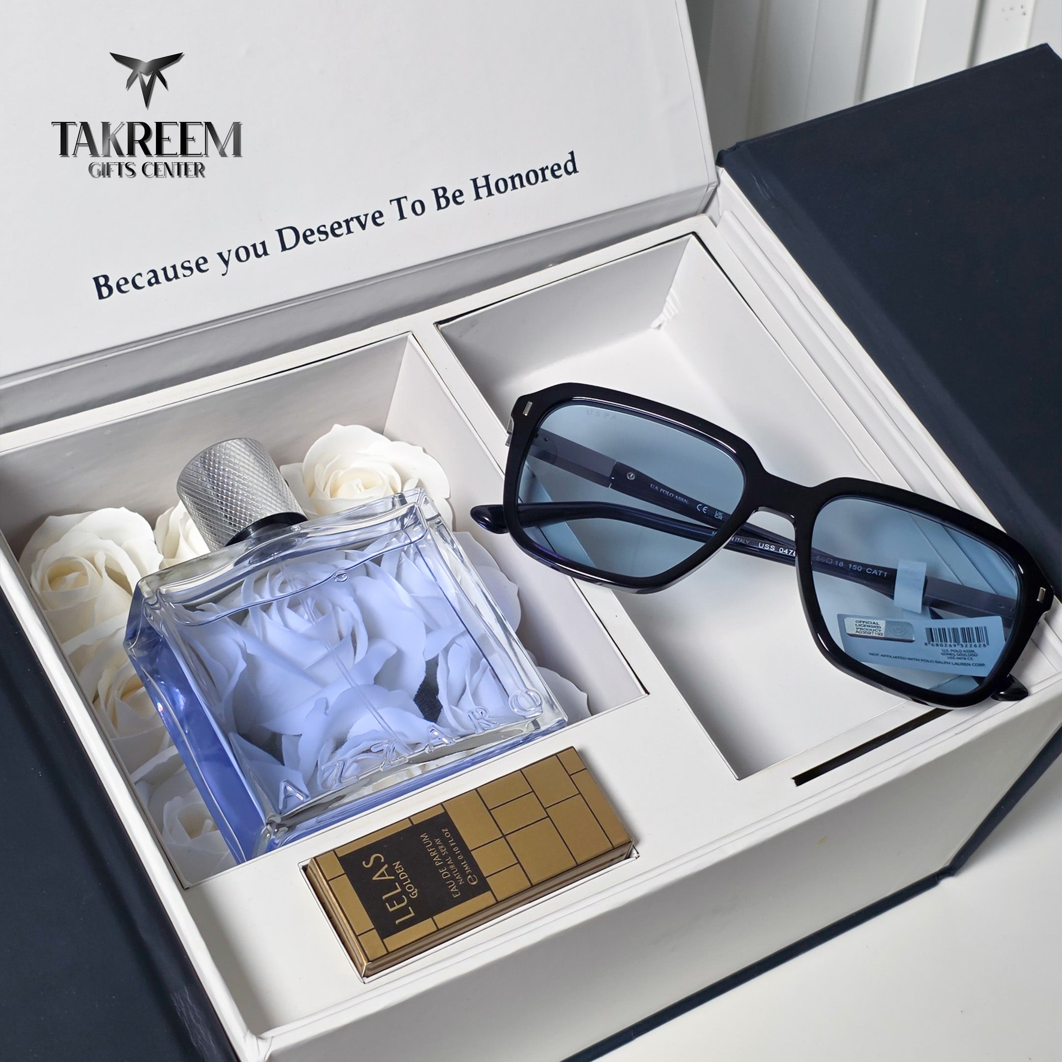 A luxury gift set from Takreem Gifts Centre featuring a pair of black rectangular sunglasses with blue lenses, a blue glass perfume bottle, and a gold chocolate bar, all arranged inside an open white box with white roses.