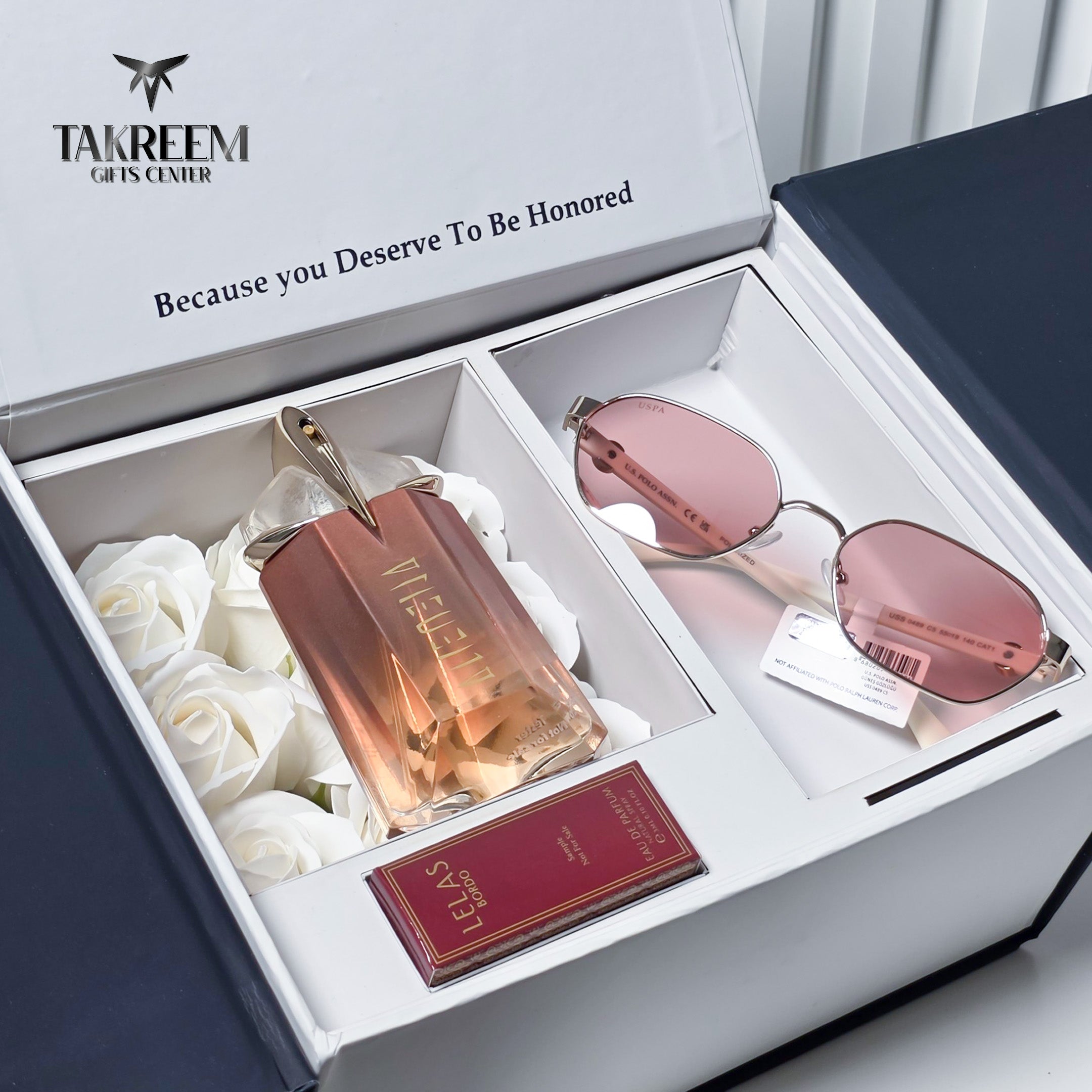 A luxury gift set from Takreem Gifts Centre in an open white box, featuring a bottle of Molecule perfume, rose gold-framed sunglasses with pink lenses, and a red card, arranged with white roses.