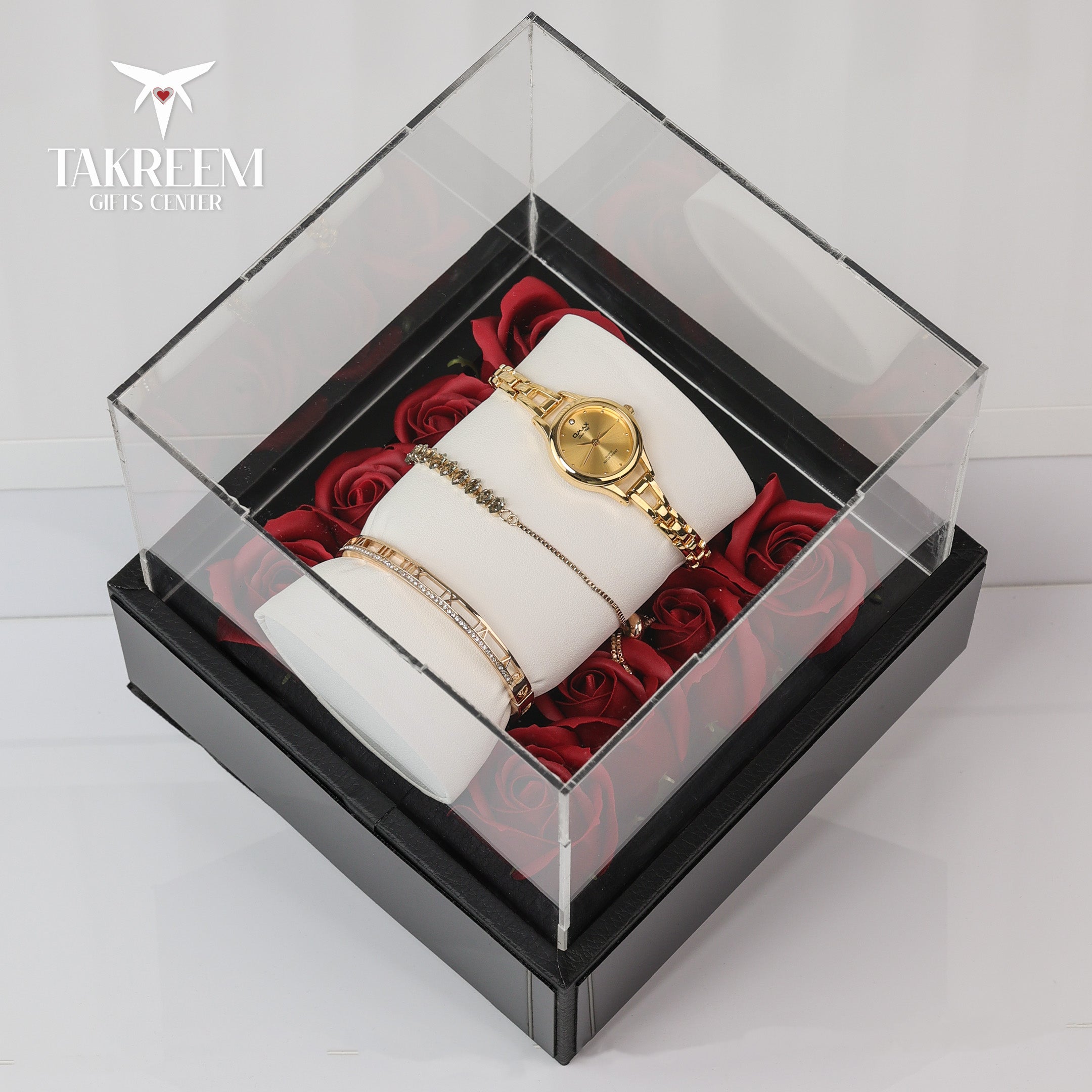 Gold watch and bracelets on a white cushion with red roses in a clear box, branded 'Takreem'.