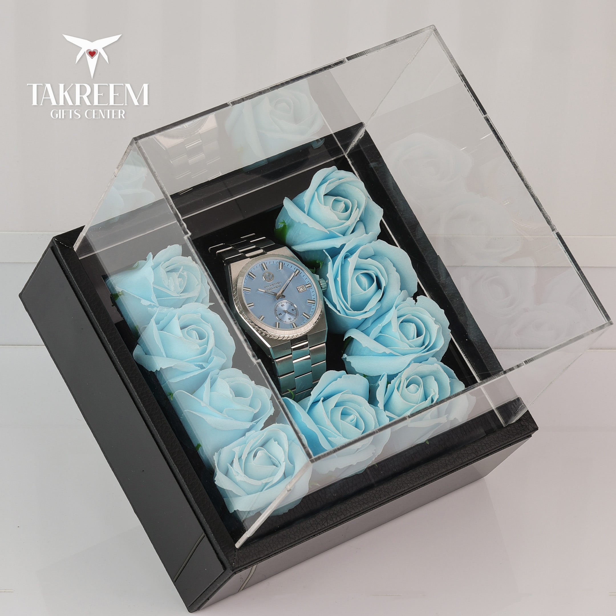Watch surrounded by blue roses in a transparent box with 'Takreem Gifts Center' branding.
