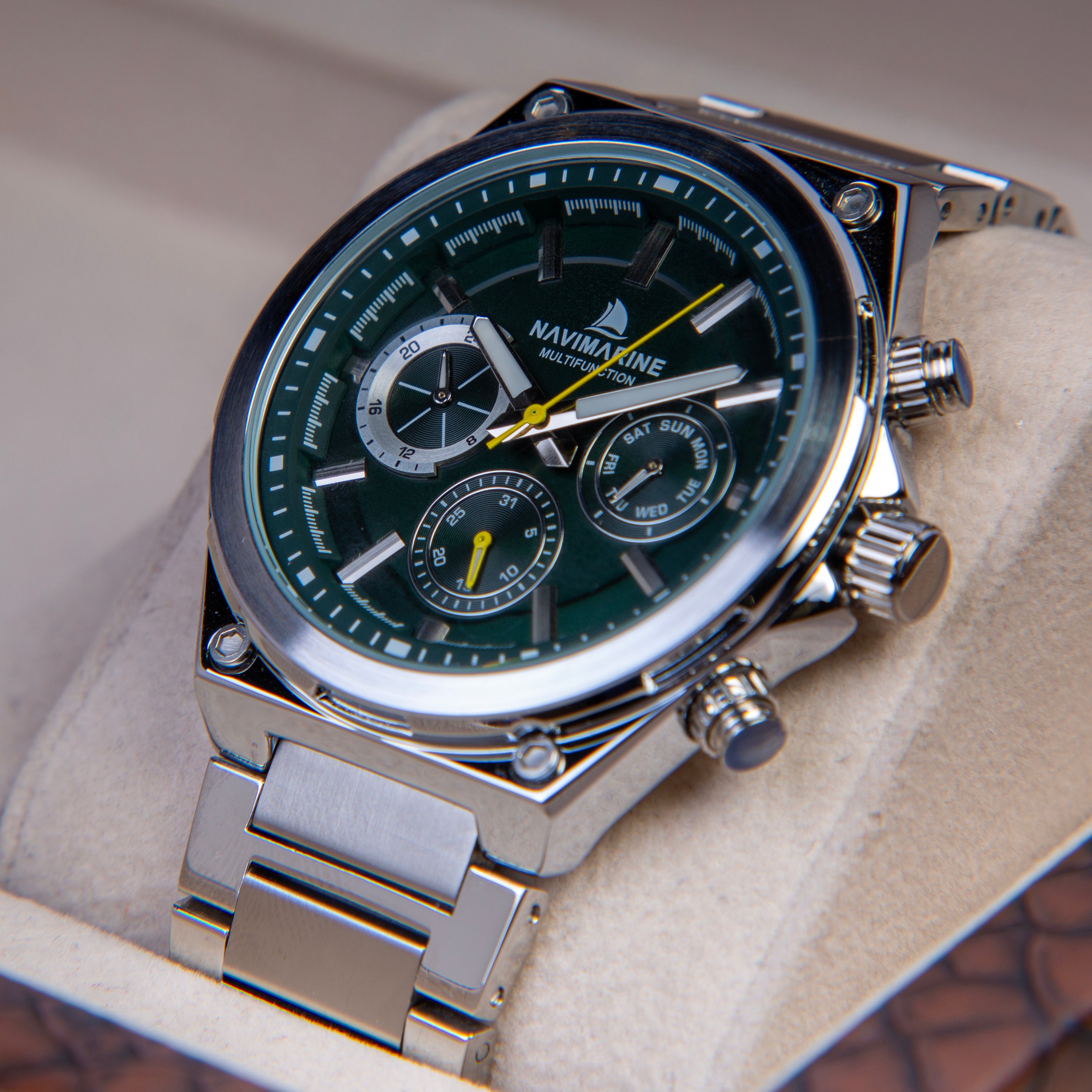 A close-up of a Navimarine stainless steel chronograph watch with a dark green dial and silver link bracelet, displayed inside a beige watch box.