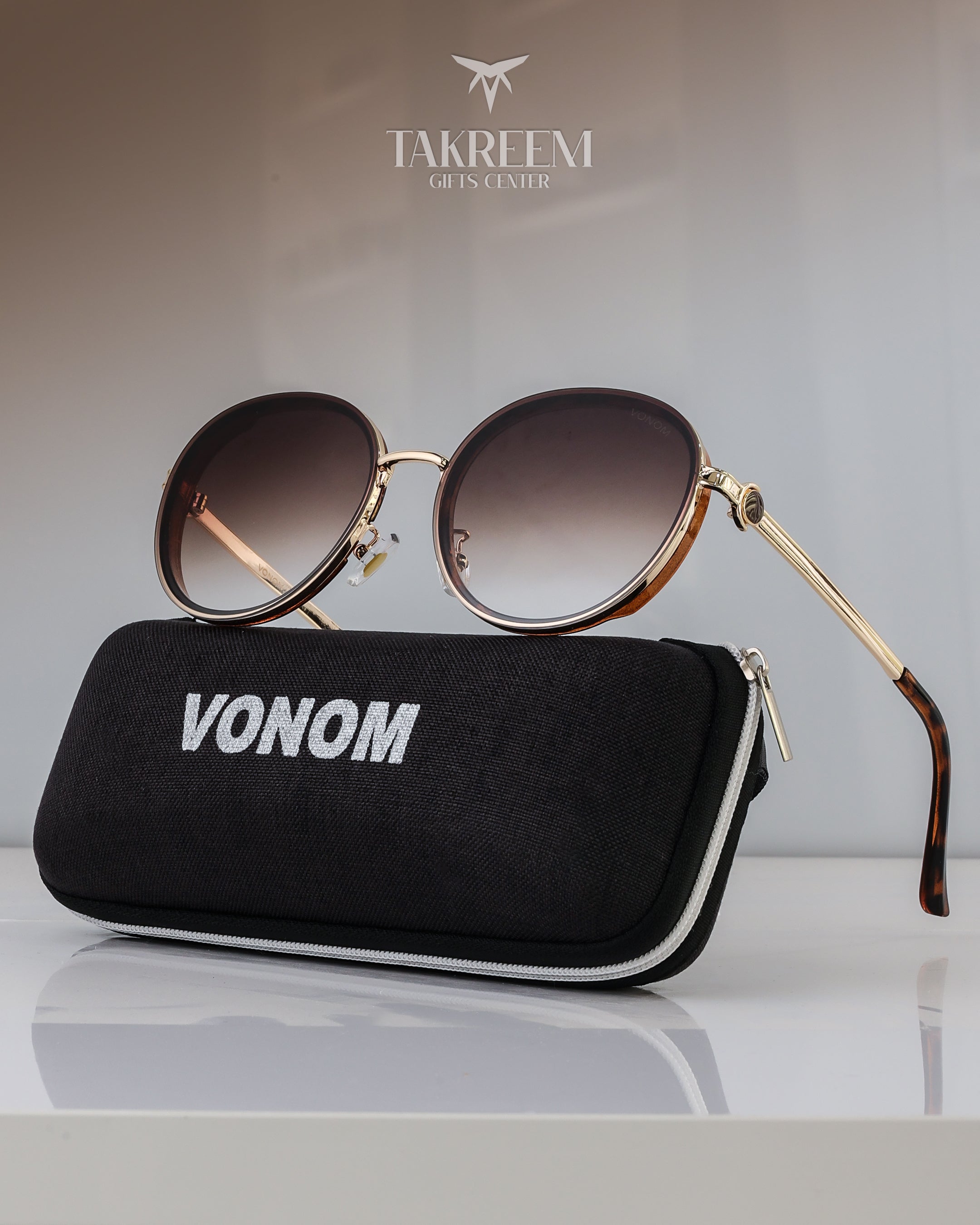 Sunglasses with gold frame and gradient lenses on a black case labeled 'Vonom' with 'Takreemi Gifts Center' logo in the background.