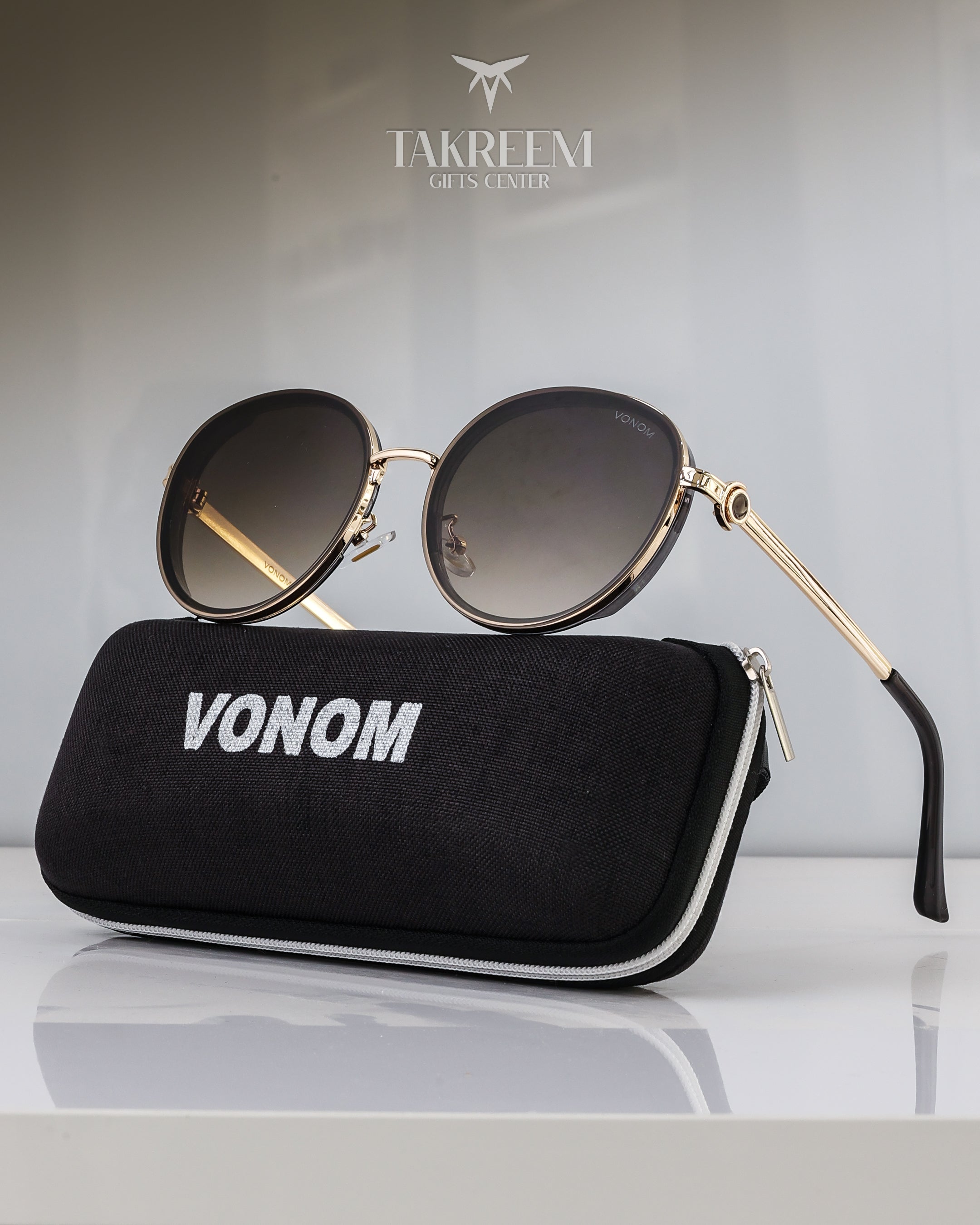 Sunglasses with gold frame and black gradient lenses on a black case with 'VONOM' branding, against a light gray background.