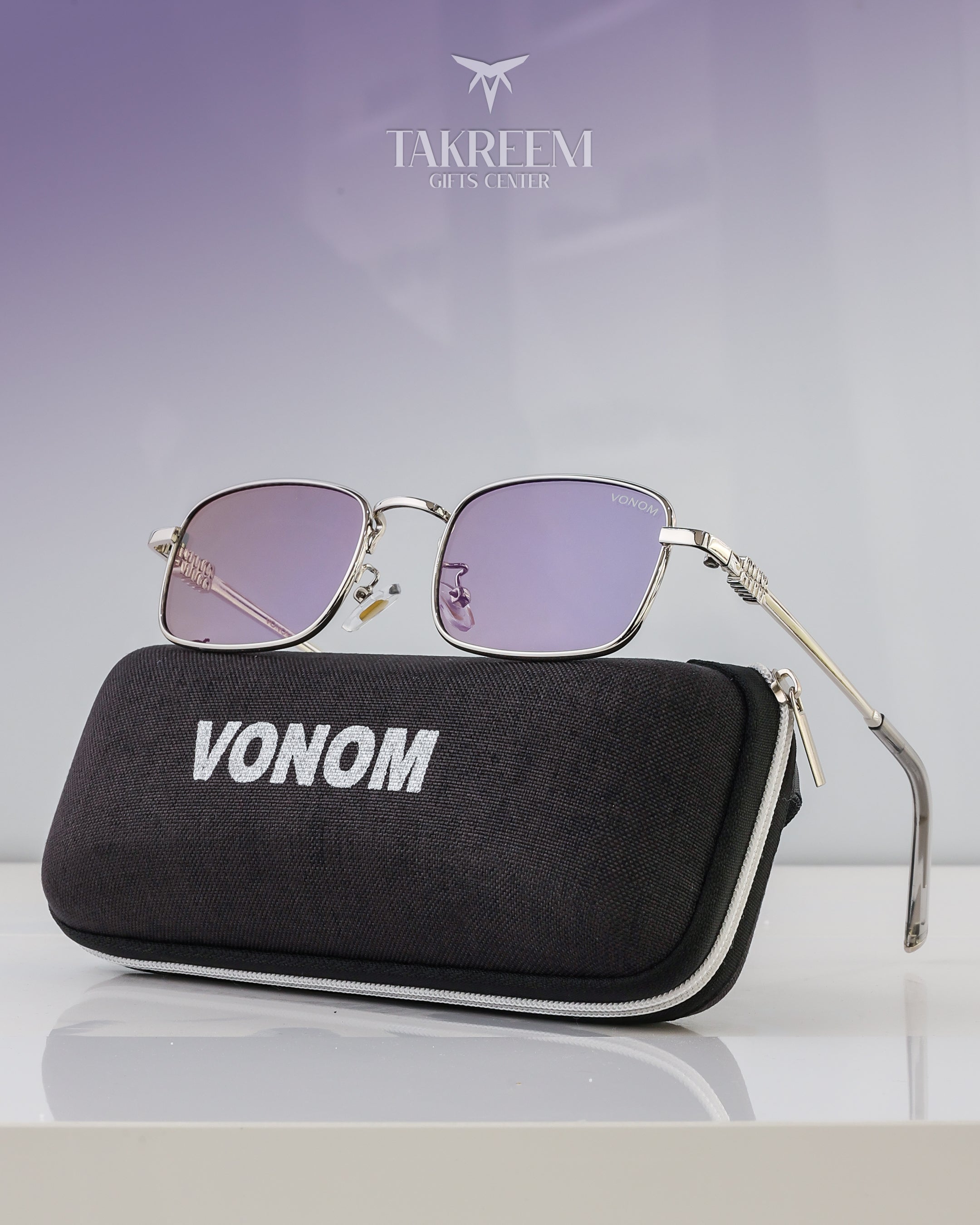 Sunglasses with a black case labeled 'VONOM' on a reflective surface with a light purple background.