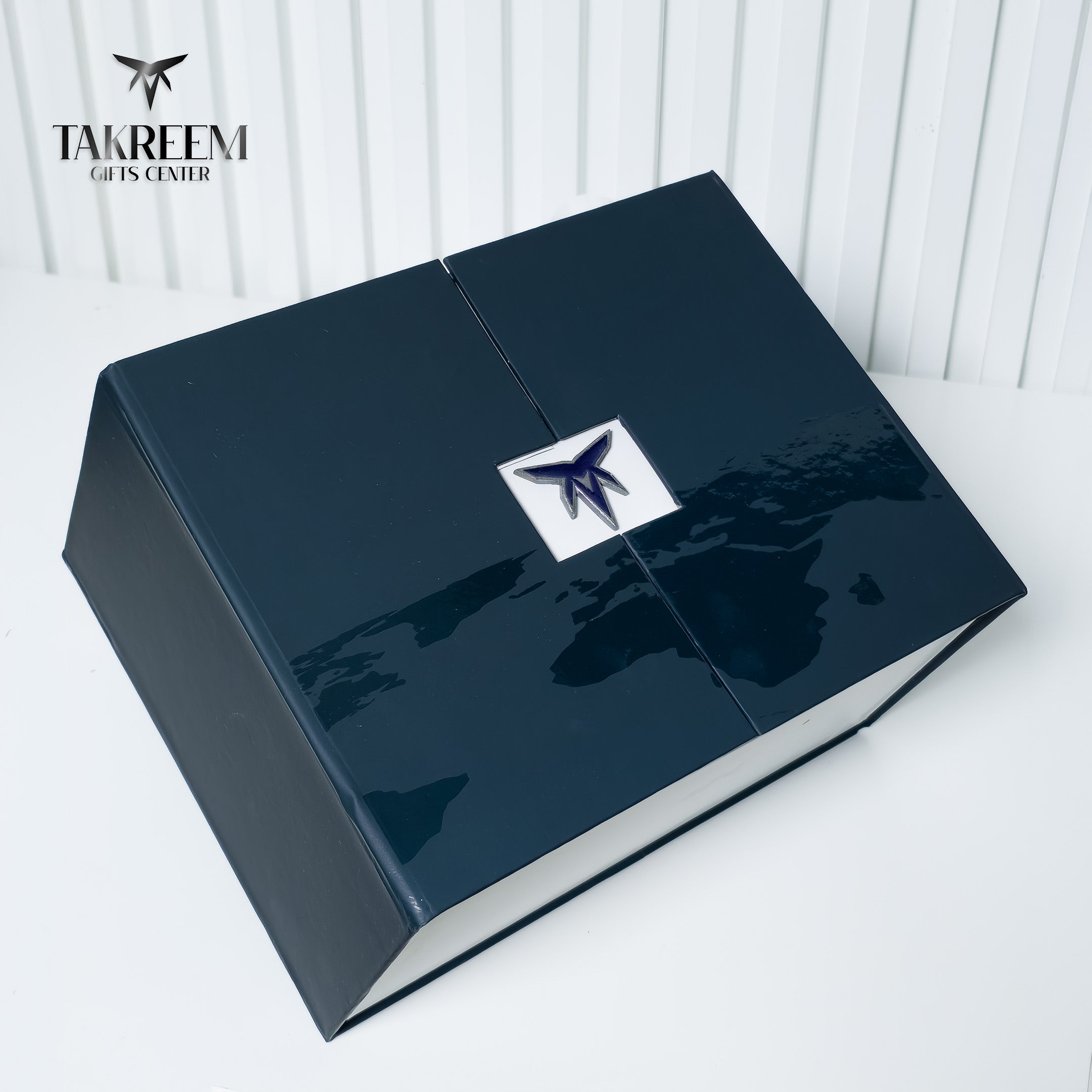 A glossy dark navy blue presentation box with a central white square featuring a blue stylized logo, photographed on a white surface against a white paneled background. The Takreem Gifts Center logo is visible in the top-left corner.