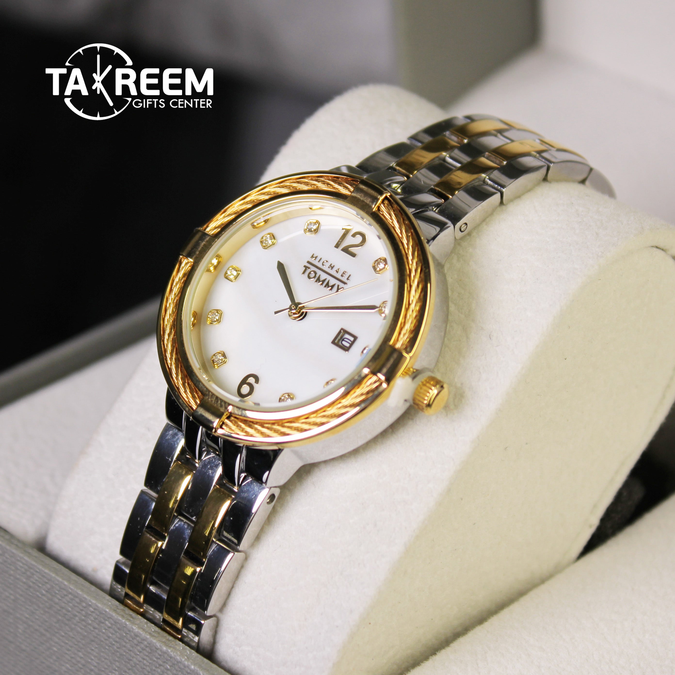 Gold and silver watch with white face on a white cushion, Taxreem Gifts Center branding visible.