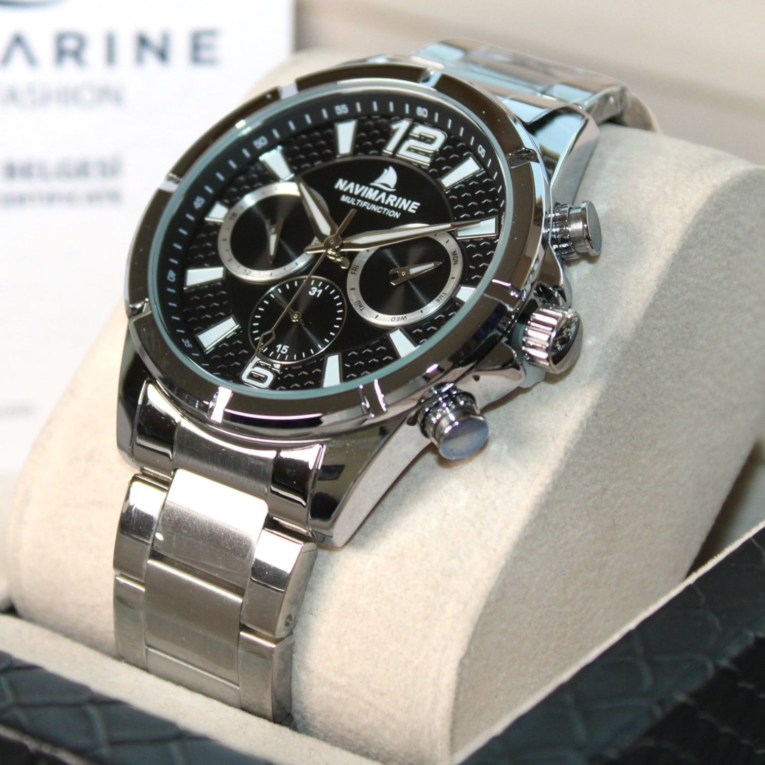 Silver wristwatch with black dial on a white cushion, brand 'Navimagine' visible.