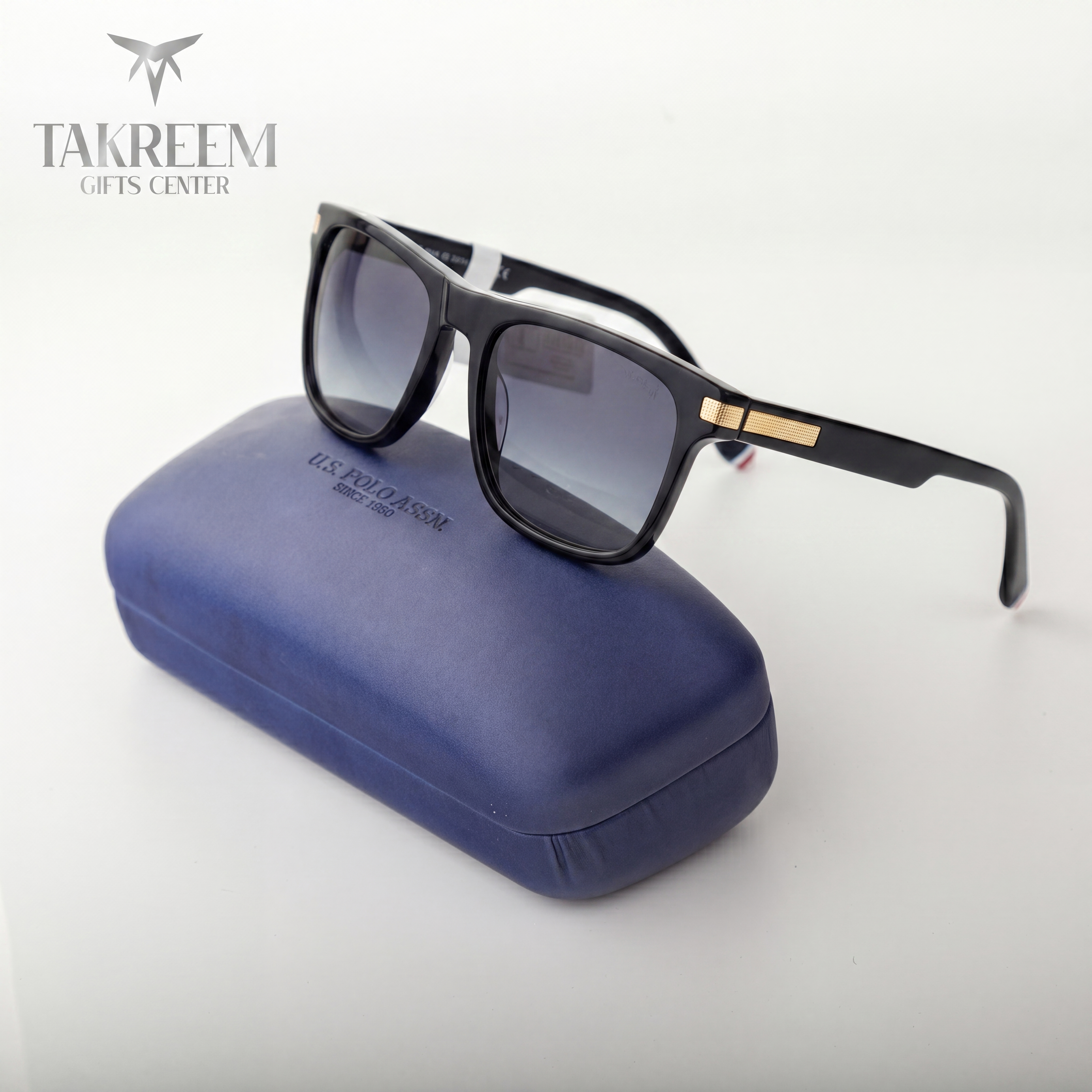 Black sunglasses with gold accents on a blue case, branded 'Takreem Gifts Center'.