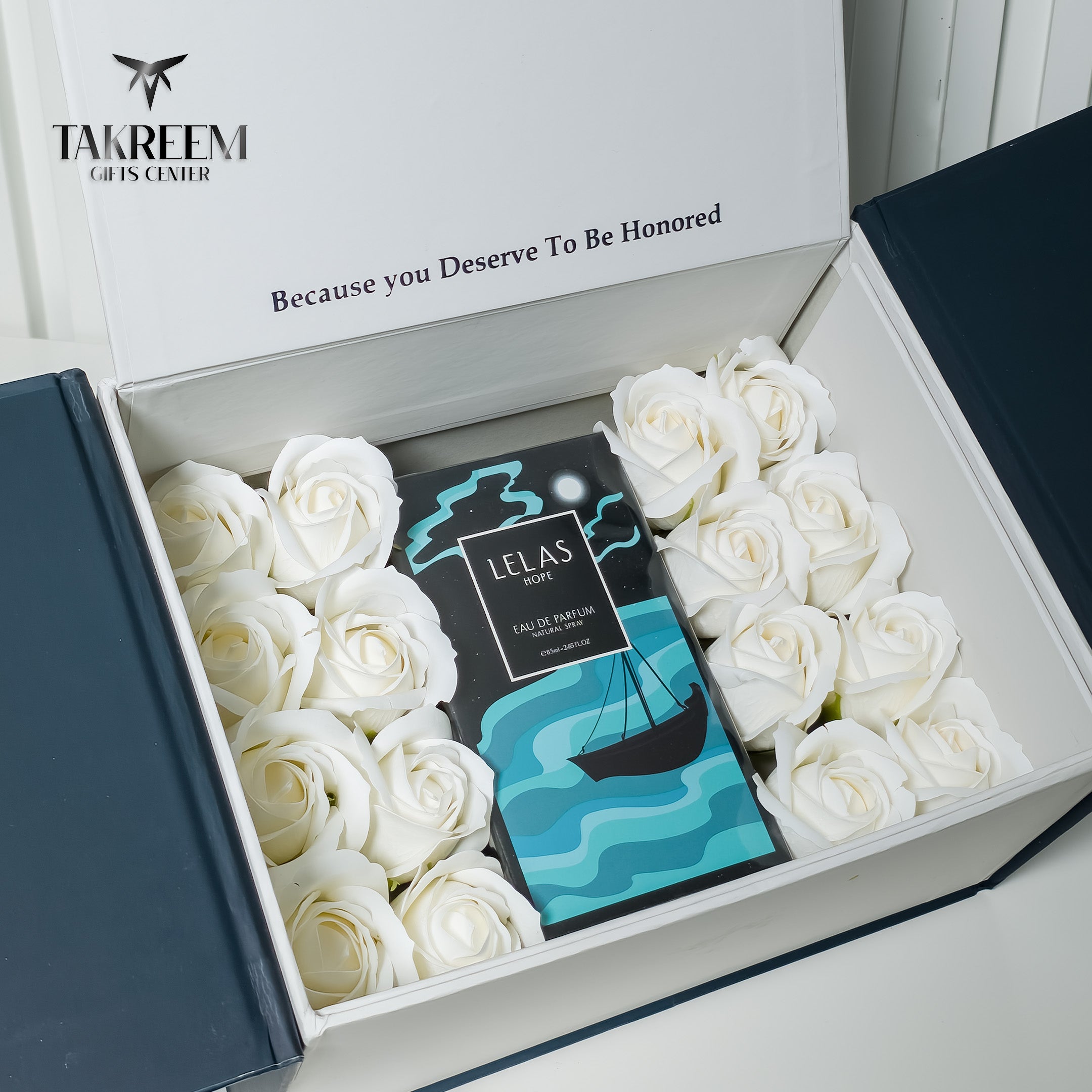 An open white gift box from Takreem Gifts Center containing white roses arranged around a central card. The card reads 'LELAS PARIS' and shows a black, teal, and white nautical design with a sailboat on waves.