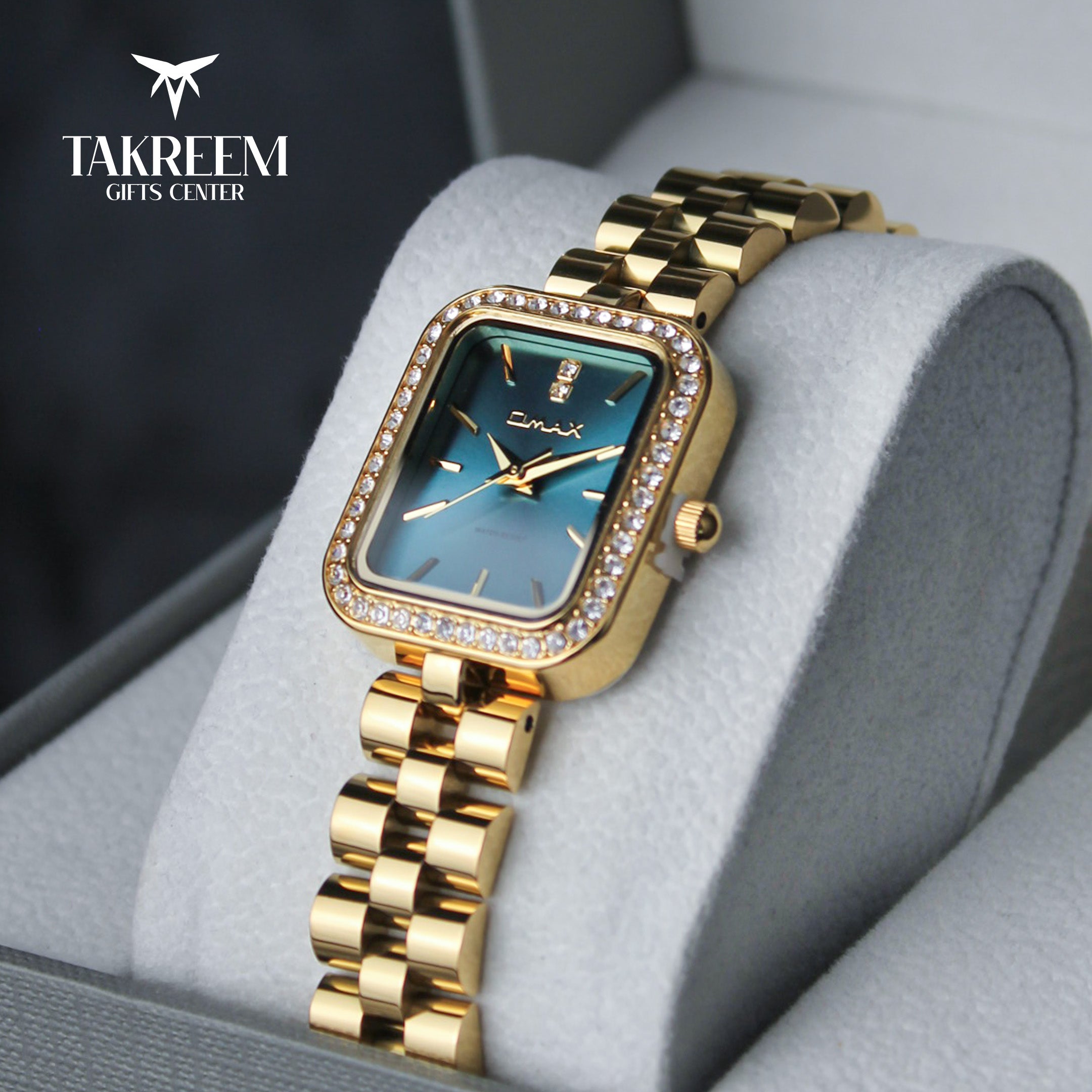 Gold wristwatch with blue face on a gray cushion, branded 'Takreem Gifts Center'.