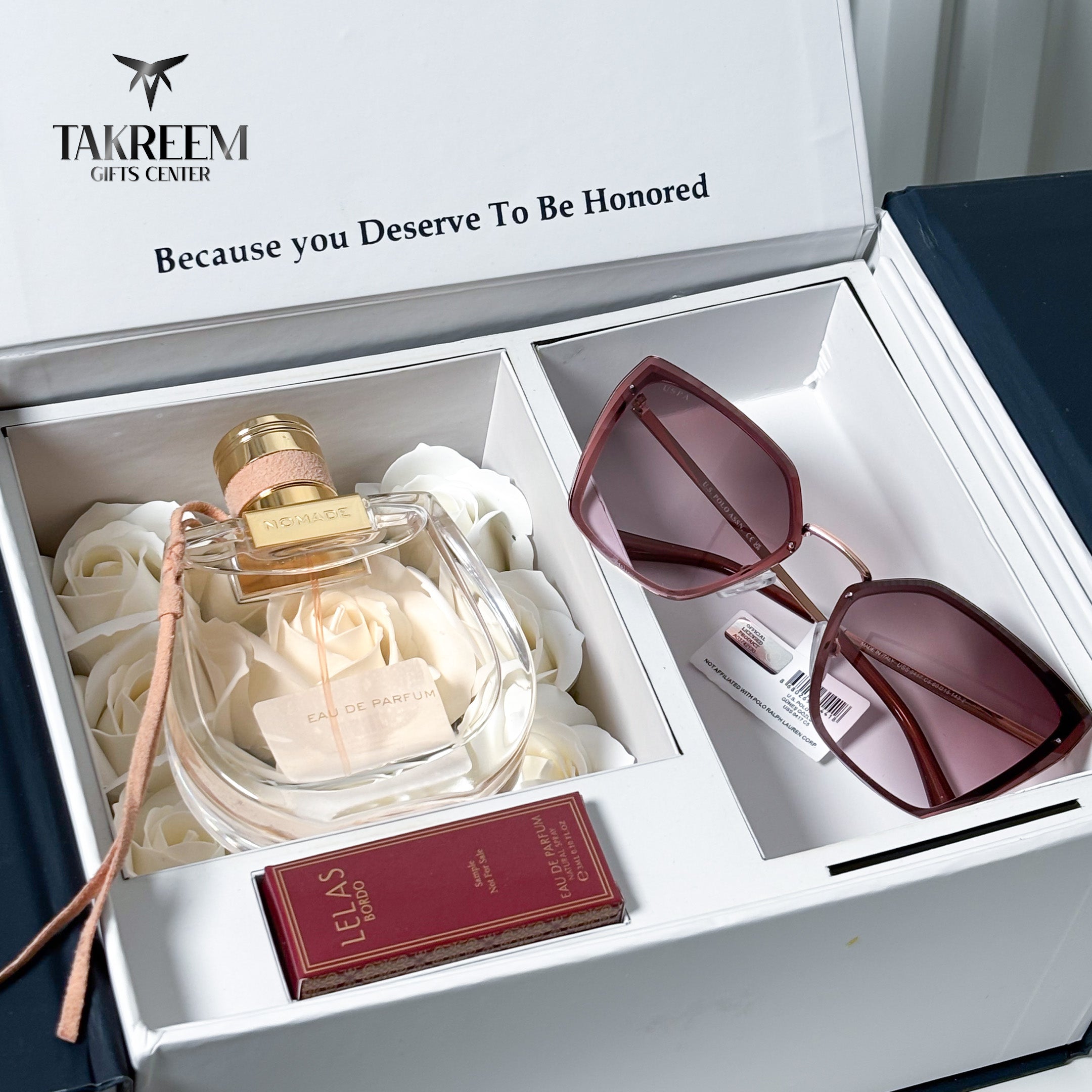 A luxury gift set from Takreem Gifts Center featuring a clear glass perfume bottle with a gold cap nestled in white roses, alongside a pair of dark pinkish-brown cat-eye sunglasses and a small red Lacoste box, all presented in a white gift box.