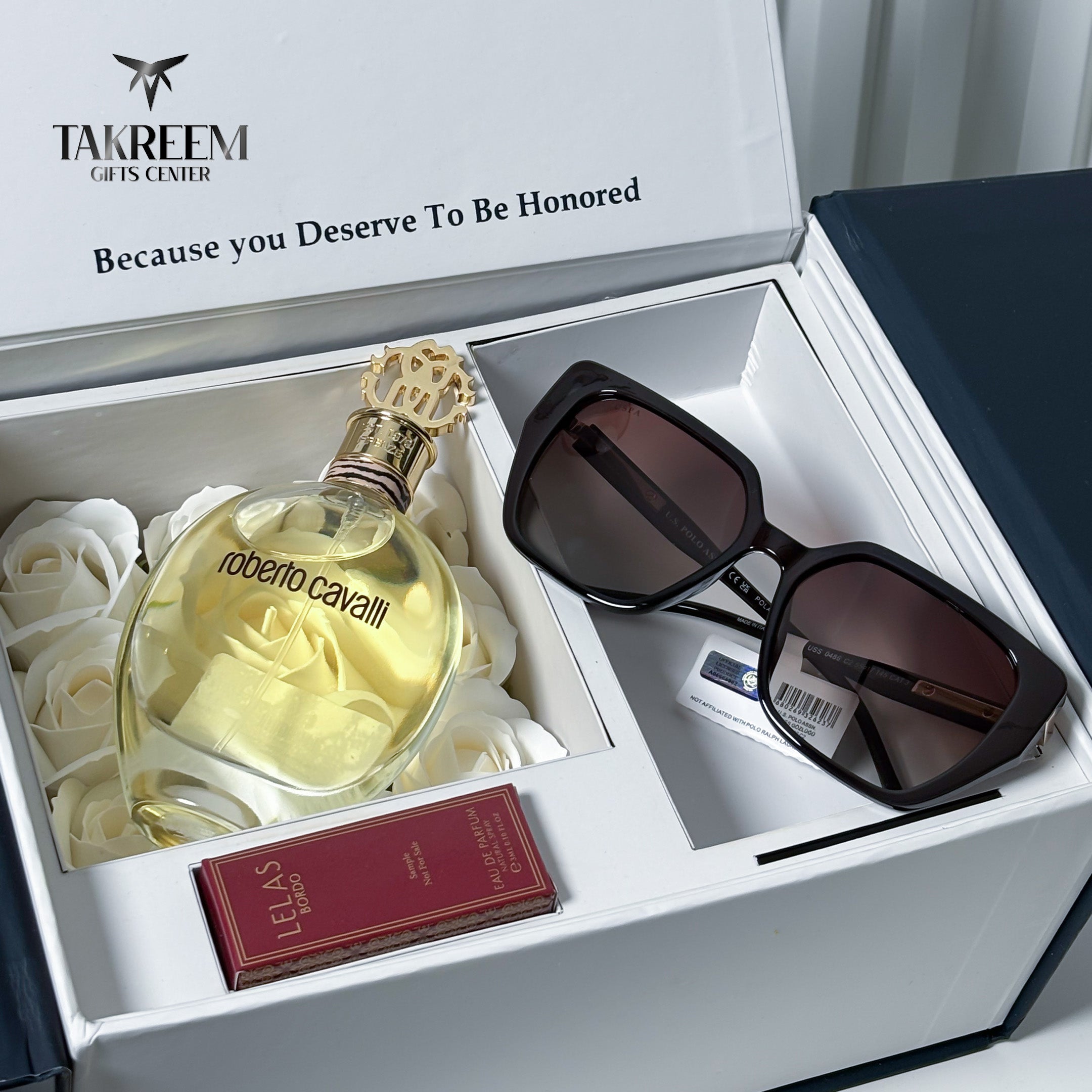 An open white Takreem Gifts Center gift box containing a Roberto Cavalli perfume bottle, black sunglasses, a small red Le Suis box, and a card, arranged on white rose petals.