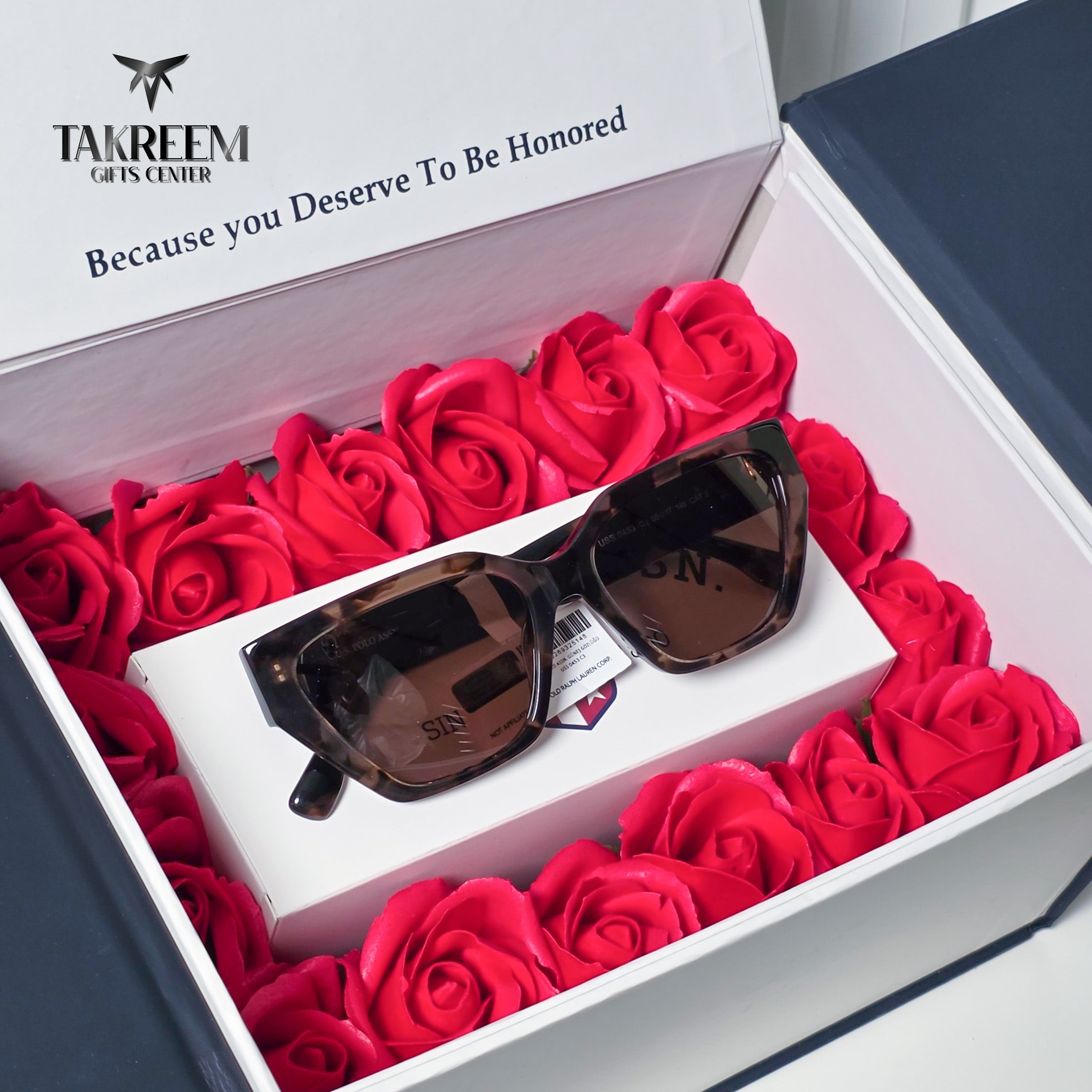 A pair of sunglasses with dark brown tortoiseshell frames and dark lenses rests on a white gift box, surrounded by a bed of vibrant red artificial roses inside an open presentation box. The inner lid features the brand name 'TAKREEM GIFTS CENTER' and the text 'Because you Deserve To Be Honored'.